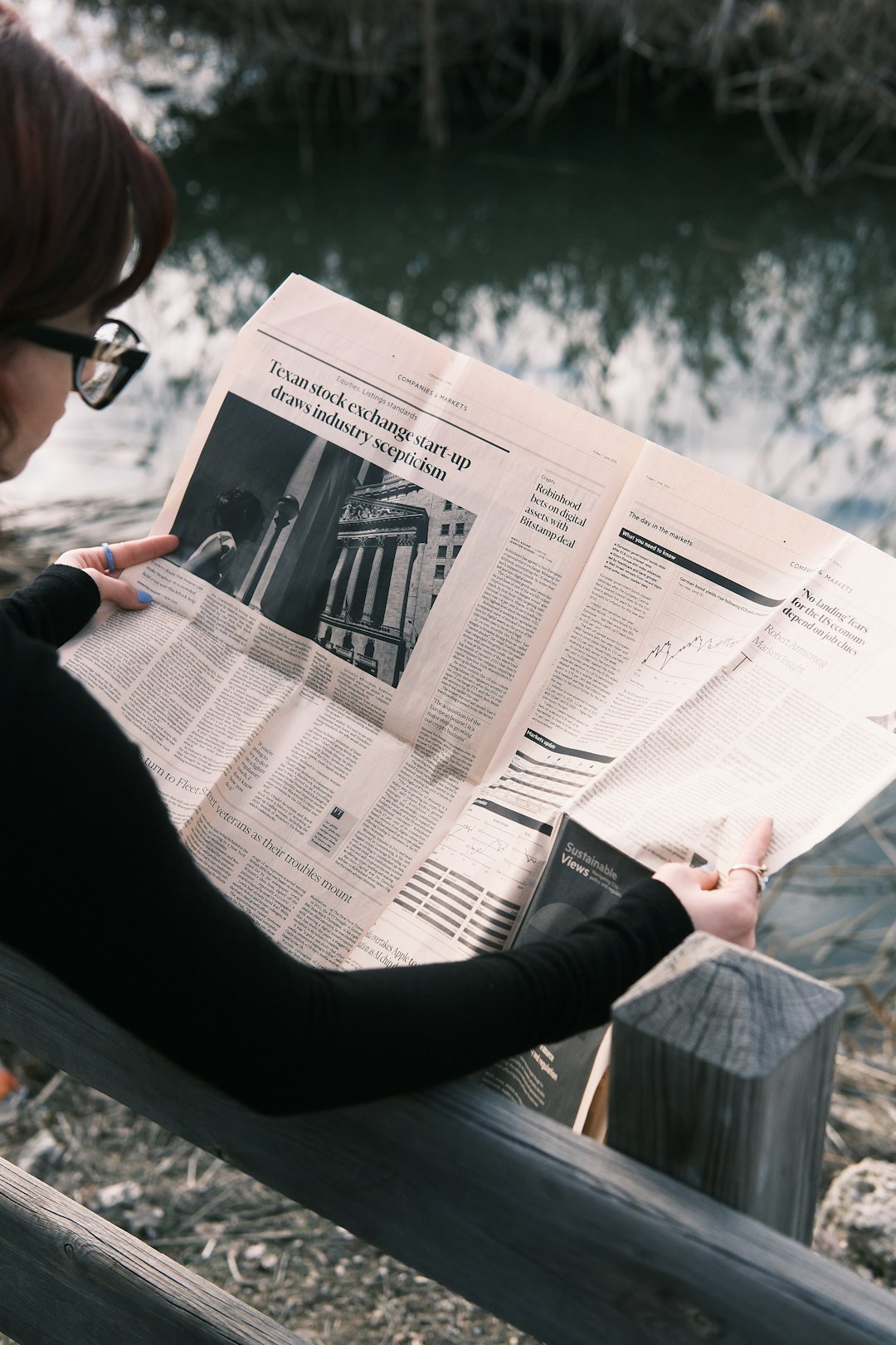 A person wearing black clothing and glasses is reading a newspaper outdoors near a calm body of water. The newspaper features articles about finance and stock markets, with visible graphs and images of financial institutions. The scene has a serene, reflective mood, with natural elements like trees and a wooden railing adding to the atmosphere. The soft lighting and muted colors create a contemplative feel.