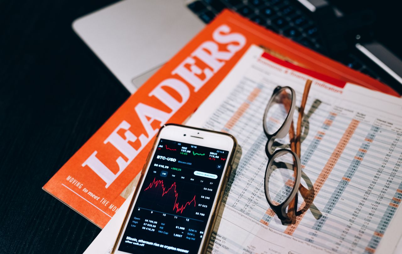 A close-up of a smartphone with cryptocurrency data next to financial reports and a magazine titled LEADERS.